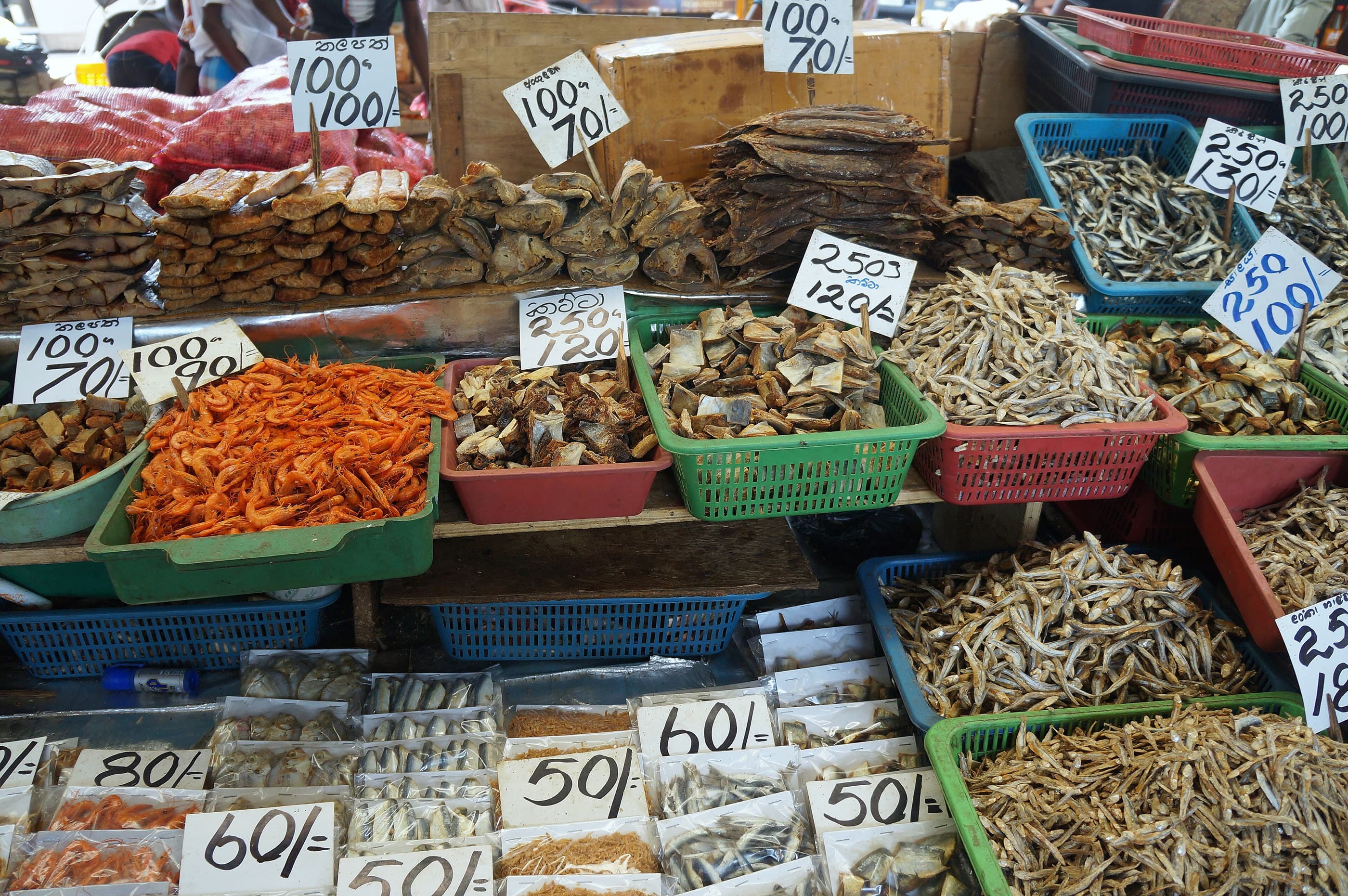 Dried fish, chillies and spices at a Colombo market stall, each tray with a handwritten price card.