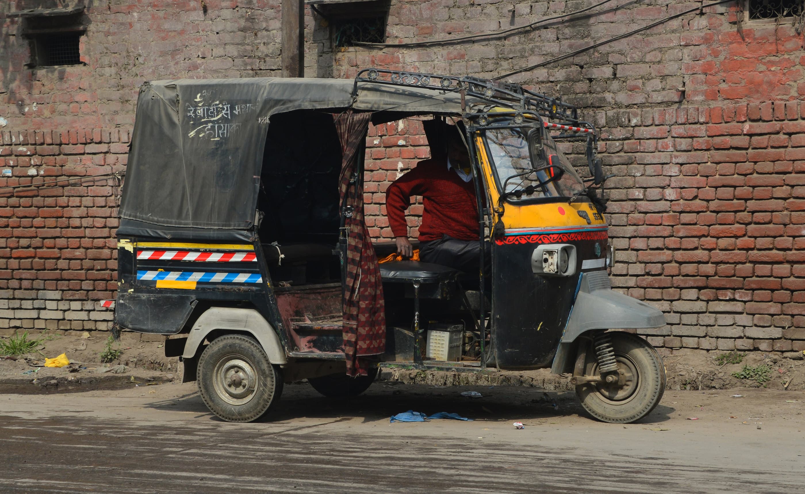 A three-wheeler parked against a brick wall, its driver at the wheel.